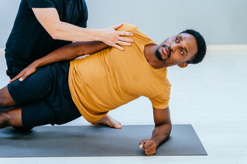 Close up of male pilates trainer helps to african man, individual training participant, do right perform. Side plank exercise, class in sport club studio led by experienced man instructor concept.
