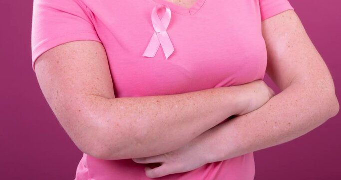 Video of midsection of caucasian woman wearing pink cancer awareness ribbon, with pink background