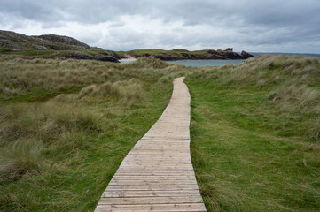 Clachtoll beach in Assynt, Scottish Highlands on a cloudy day with dramatic sky. Wooden boardwalk leading through grass to beach, sea and rocks visible, no people.