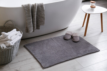 Soft grey mat with slippers on floor near tub in bathroom. Interior design