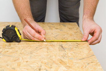 Young adult man hands measuring osb board with measure tape and marking precise size with pencil. Closeup. Front view. Preparing material for new floor or wall. Home repair work. Renovation process.