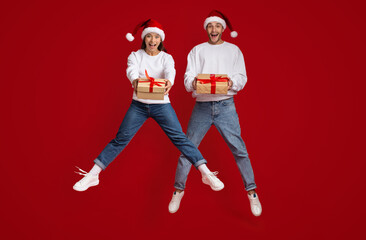 Excited Millennial Couple Wearing Santa Hats Jumping With Christmas Gifts In Hands