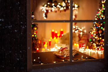 Christmas Decorated Room Interior with Garland Lights, Candles and Fir Tree. Winter Frosted Window Frame. Xmas Holiday Gingerbread Cookies for Santa at Table