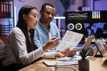 Male and female coworkers preparing financial report during evening time at office. Asian woman and african man using modern laptop and tablet for working process.