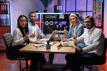 Four multi ethnic office workers showing thumb up and smiling on camera during business meeting. Coworkers using charts, graphs and technology while discussing common project.