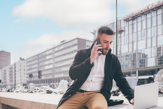 Young caucasian bearded contemporary businessman freelancer remote working outdoor city talking smartphone using computer