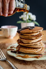 Hand pouring maple syrup on a healthy buckwheat pancake stack topped with chocolate and halvah on a decorative plate. Easy to make gluten free morning breakfast, brunch. Delicious golden pancake heap.