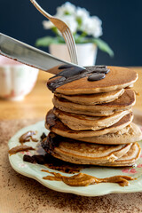 Eating healthy buckwheat pancakes with a knife and fork. Delicious stack of golden pancakes with chocolate, maple syrup, and halvah spread on a decorative plate. Easy to make gluten free breakfast.