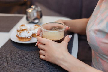 
The girl has breakfast and drinks cappuccino from a glass mug, eats cream cakes. Breakfast and coffee concept