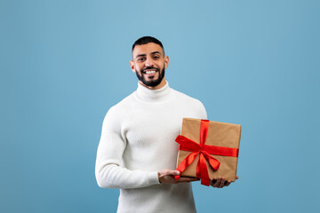 Best gift for you. Happy young arab man holding wrapped present box, smiling to camera, standing over blue background