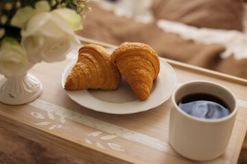 The atmosphere of a romantic morning, coffee in bed. Flowers in a vase, french croissants and a cup of coffee on a wooden tray on a silk bed. Home interior. Lifestyle