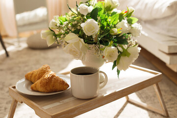 The atmosphere of a romantic morning, coffee in bed. Flowers in a vase, french croissants and a cup of coffee on a wooden tray on a silk bed. Home interior. Lifestyle