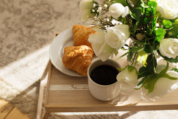 The atmosphere of a romantic morning, coffee in bed. Flowers in a vase, french croissants and a cup of coffee on a wooden tray on a silk bed. Home interior. Lifestyle