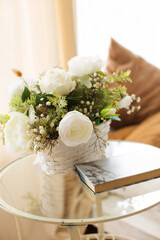 
White artificial roses in a basket on the coffee table, next to a book for reading. Interior decoration in pastel colors