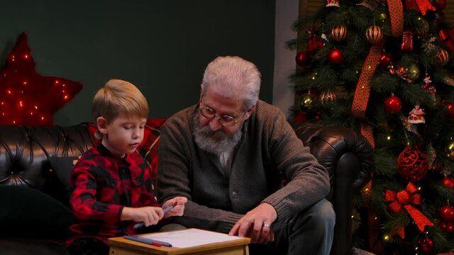 Little boy draws a festive drawing with felt-tip pens. Grandfather and grandson sit on a sofa in a decorated room near a glowing Christmas tree. Family vacation concept. Close up. Slow motion.