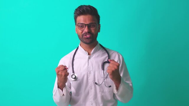 indian bearded cardiologist man with eyes glasses in white medical coat with stethoscope in blue studio background telling a good news