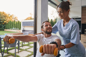 Adorable Caucasian male holding dumbbell in hands while looking to the female physiotherapist in the room indoors