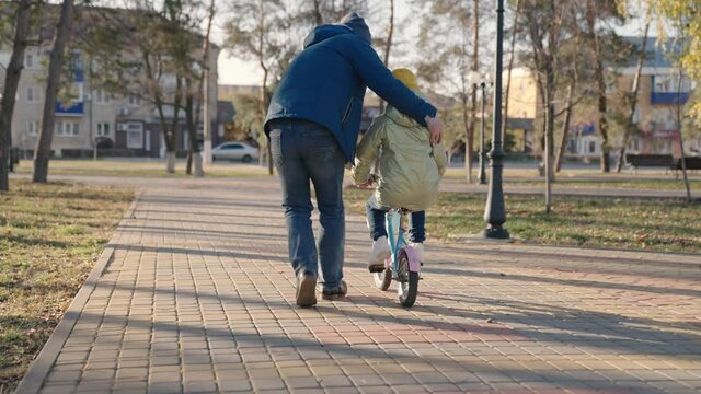 father teaches little kid to ride bike on road in city park, child spins pedals and wheels, happy family, dad helps the girl to ride daughter, play in autumn park, parent and baby have fun together