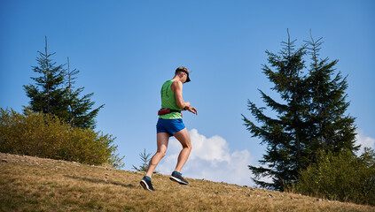 Trail runner running up to meadow outdoors under blue sky and looking at his watch. Jogging in open nature space in sunshine warm day. Concept of sport, training and active leisure.