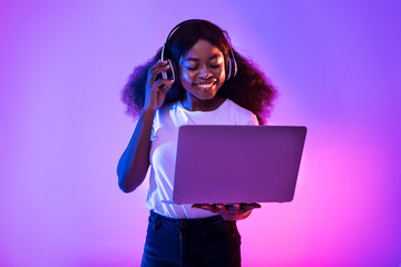 Young black woman having video call, using laptop computer, wearing wireless headphones in neon...