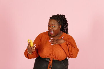 Wondered young black plus size body positive woman with dreadlocks in orange top holds smartphone standing on light pink background in studio closeup - 466753509