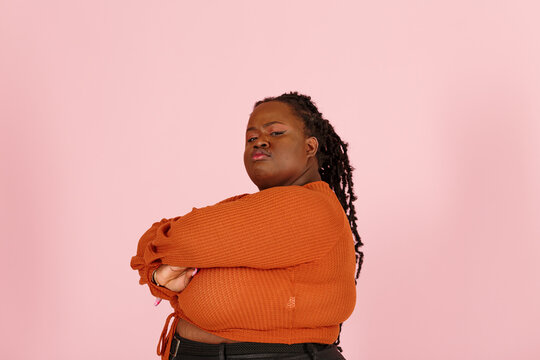 Critical young black plus size body positive woman with dreadlocks in orange top looks at camera crossing arms on pink background in studio