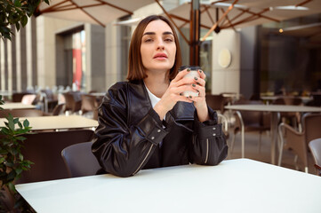 Lifestyle portrait of well-groomed brunette, European woman, with cardboard coffee mug looking at camera while resting in summer terrace of outside cafeteria restaurant, enjoying autumn cool weather