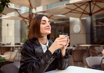 Delighted attractive young woman with closed eyes of pleasure, cutely smiles enjoying coffee break, resting in outside cafe, holding takeaway paper cup with hot drink. Freedom, freelancer, lifestyle.