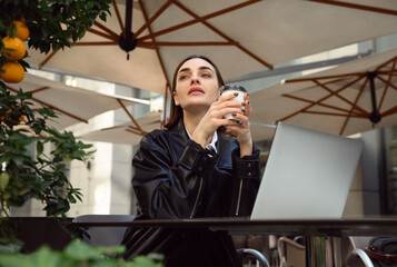 Cheerful pretty woman with stylish haircut, in black eco leather jacket holding a takeaway paper cup of delicious hot drink and enjoying coffee break in outside cafe during online work at sunny day