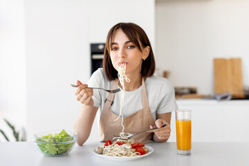 Happy lady eating spaghetti and vegetable salad, enjoying tasty lunch, sitting at table and looking at camera in kitchen