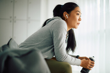 Pensive Asian sportswoman listens music over earphones in locker room at gym.
