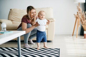 Happy mother assists her small son in taking first steps at home.