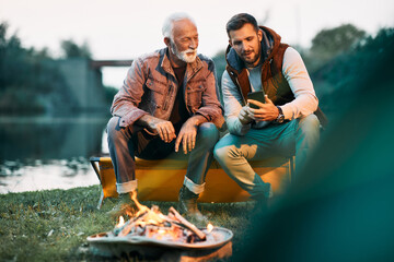 Mid adult man and his senior father use smart phone by campfire in nature. - obrazy, fototapety, plakaty