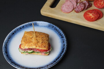 sandwich sandwich of white square bun with vegetables tomato cucumber sausage ham on a blue plate. in the background is a tray board with ingredients vegetables tomatoes on a black background. 