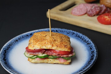 sandwich sandwich of white square bun with vegetables tomato cucumber sausage ham on a blue plate. in the background is a tray board with ingredients vegetables tomatoes on a black background. 