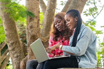 two female african students sitting on a park bench using a laptop to check something online