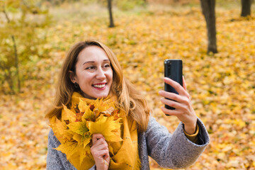 Smiling young woman holds autumn yellow leaves takes selfie in the autumn park. Autumn mood. Close-up.