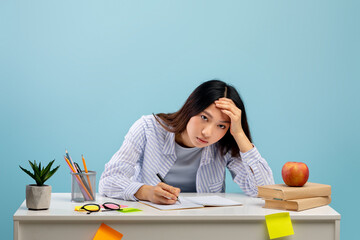 Tiresome task. Exhausted asian lady sitting at table, writing in notebook, studying over blue background