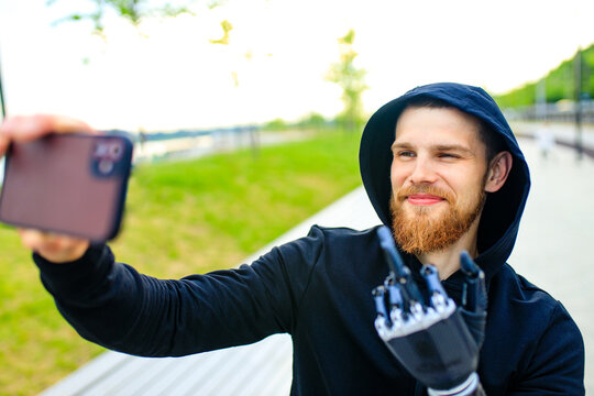 young bearded man with metal arm prosthetic outdoor taking selfie photos on smartphone camera