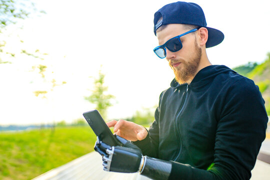 young bearded man with metal arm prosthetic outdoor looking at screen of smartphone