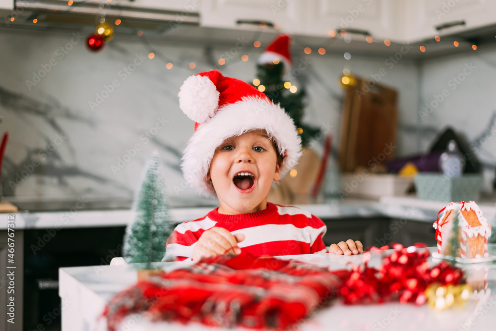 Wall mural Funny boy child 3 y.o. in a Santa hat is sitting in the kitchen with a decor for Christmas. Holidays, Traditions, baby food and health concept.