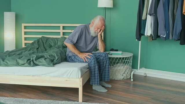 Tired Elderly Man Has Acute Headache While Sitting on ahe Bed At Home. Virus, Headache, Migraine, Anxiety. Seniors, Dizziness, High Blood Pressure, Health Care and Retirement Insurance.