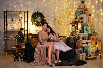 Obraz na płótnie Canvas Christmas and new year concept - beautiful mother with her cute daughter sitting in decorated room with heap of christmas gifts
