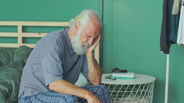 Tired Elderly Man Has Acute Headache While Sitting on ahe Bed At Home. Virus, Headache, Migraine, Anxiety. Seniors, Dizziness, High Blood Pressure, Health Care and Retirement Insurance.