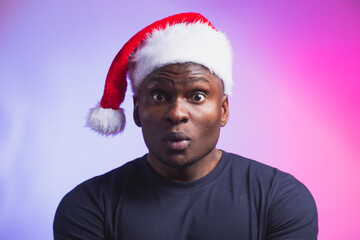 Portrait of surprised african american man in santa hat and casual t-shirt on colourful background, christmas time. Winter holidays and emotional people concept.