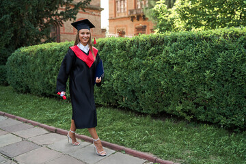 Pretty graduate girl in graduations gown walking with diploma in campus