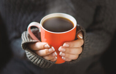 Beautiful hands of a young girl, with a red cup, the concept of a winter morning. A girl in a warm knitted cardigan holds a red mug with a hot drink in her hands. The concept of comfort, warmth.
