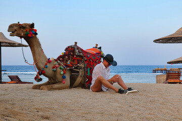 A beautiful woman in a big hat, with a phone in her hands sits near a camel.