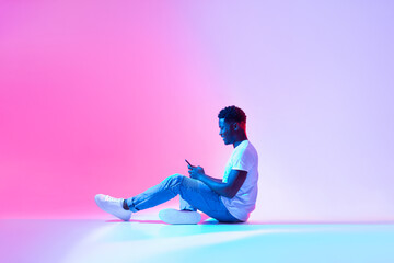 Cool mobile app. Side view of happy black man using modern smartphone in neon light, copy space