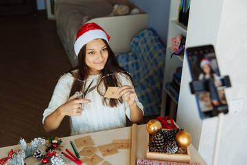 a smiling blogger girl in a Christmas hat makes an advent calendar and takes pictures on her phone